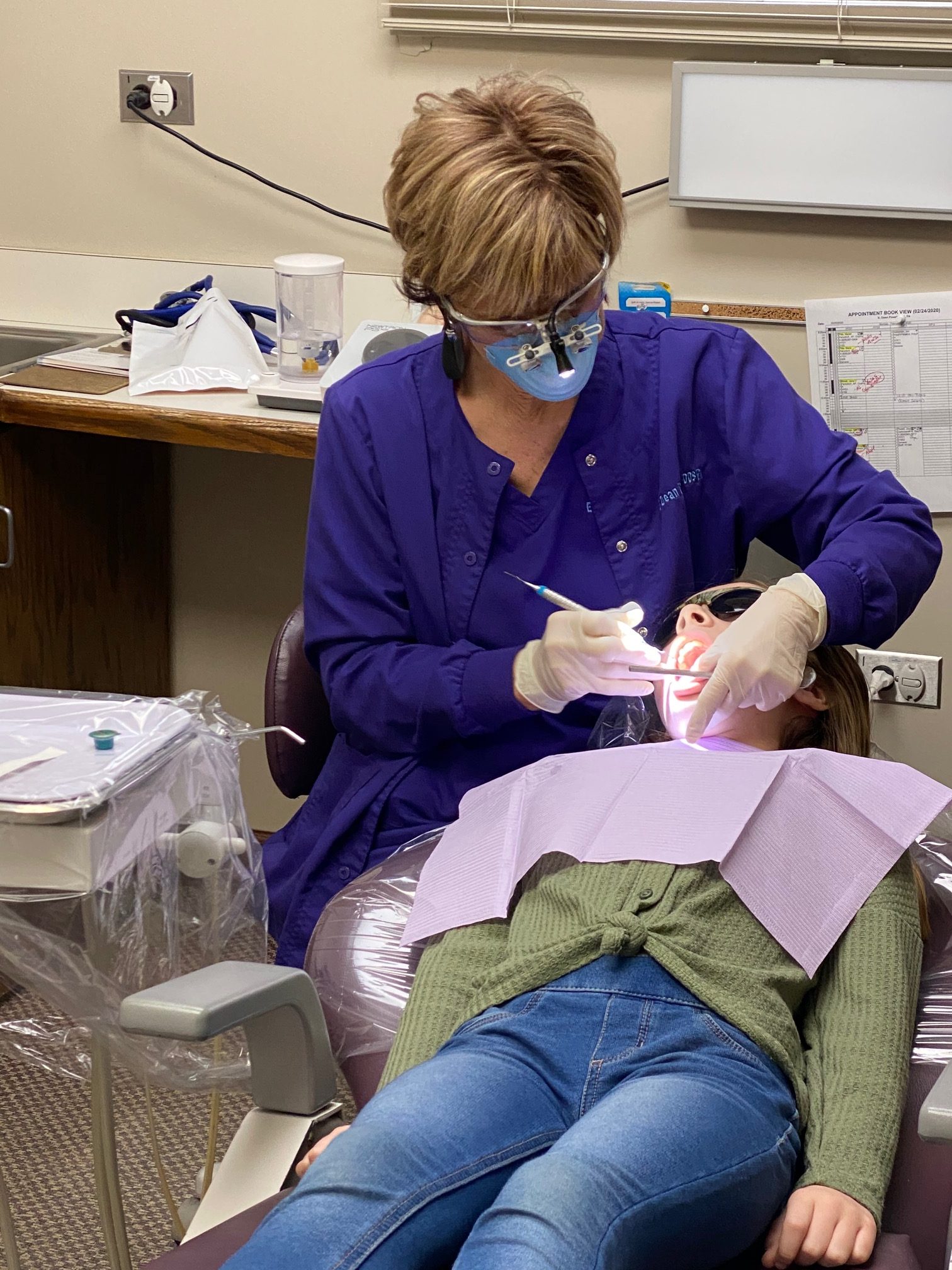 Susan McLain, Senior Hygienist, is cleaning a patient's teeth and wearing protective glasses, face mask, and gloves.