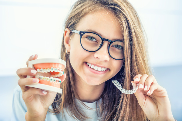 invisalign-vs-braces A young girl holds teeth molds with traditional braces in one hand with different colored bands around the brackets and an Invisalign aligner in the other.