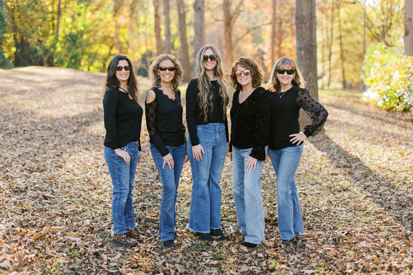 The team in black sunglasses and black tops while smiling and standing in the woods wearing blue jeans.