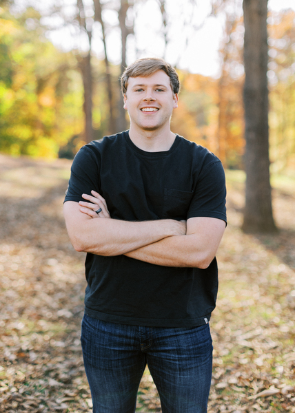 Dr H smiling while standing in the woods dressed in a black top and blue jeans